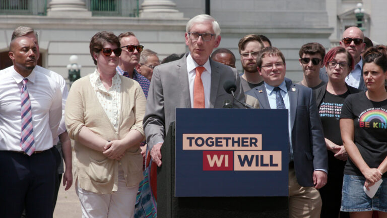 Tony Evers stands behind a podium affixed with a sign reading Together WI Will and speaks into a microphone, with other people standing behind and to either side of him, with a marble masonry building with  the bases of pillars in the background.