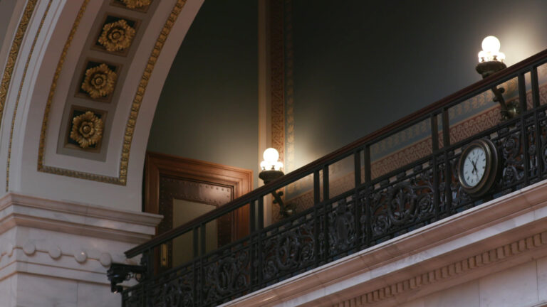 A clock is mounted to iron filigree decoration on a railing for a gallery seating area in the upper portion of a room with marble masonry and illuminated electric brass wall sconce light fixtures. 