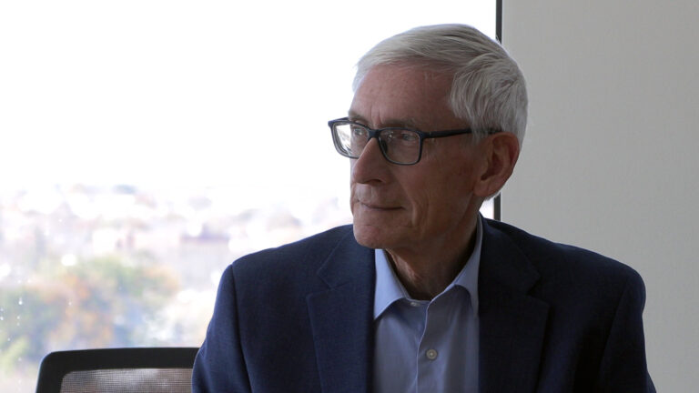 Tony Evers sits and listens in a room with sunlight illuminating a window in the background.