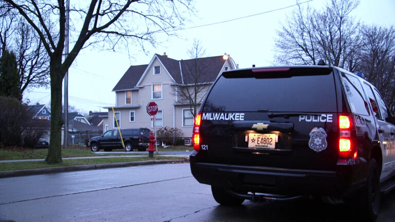 An SUV with the words Milwaukee Police and a badge logo on its back and its brake lights illuminated is parked near an intersection, with a stop sign, parked vehicles, houses and leafless trees in the background.