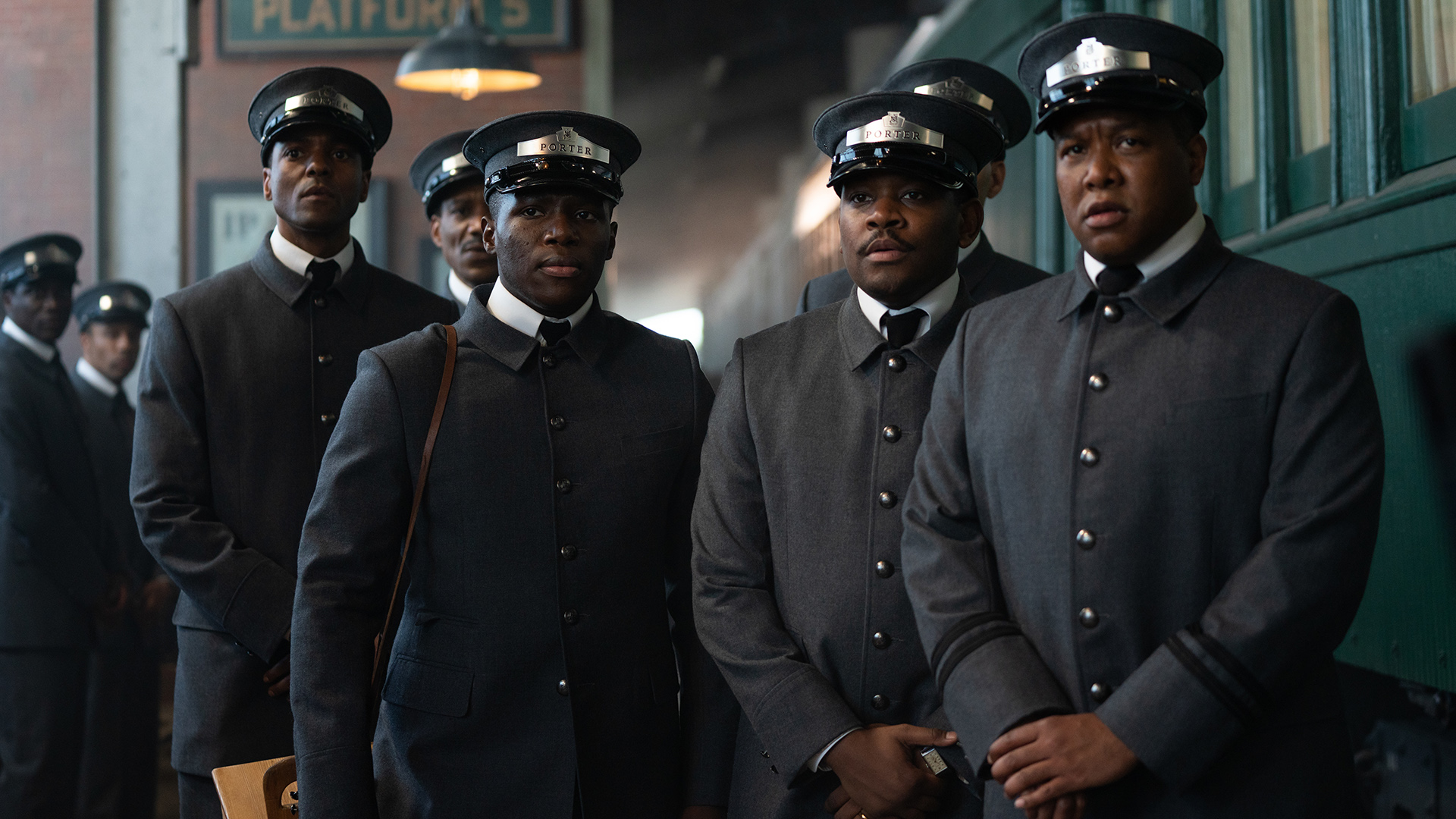 A group of Black men wearing dark gray uniforms and black caps stare at the camera with serious expressions.
