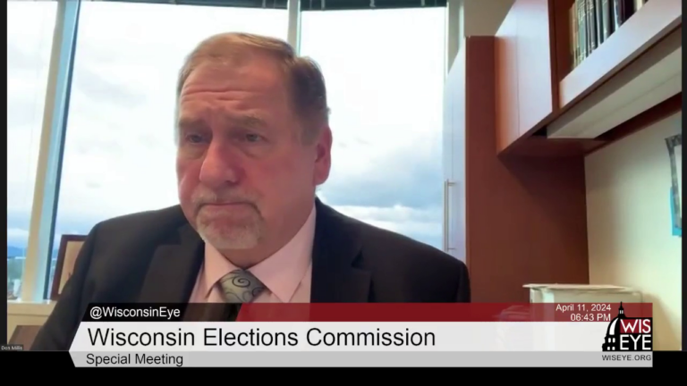 A video still image shows Don Millis sitting in an office with wood cabinets on one side and large plate-glass windows in the background showing a cloudy sky, with a video graphic at bottom including the text Wisconsin Elections Commission and Special Meeting.