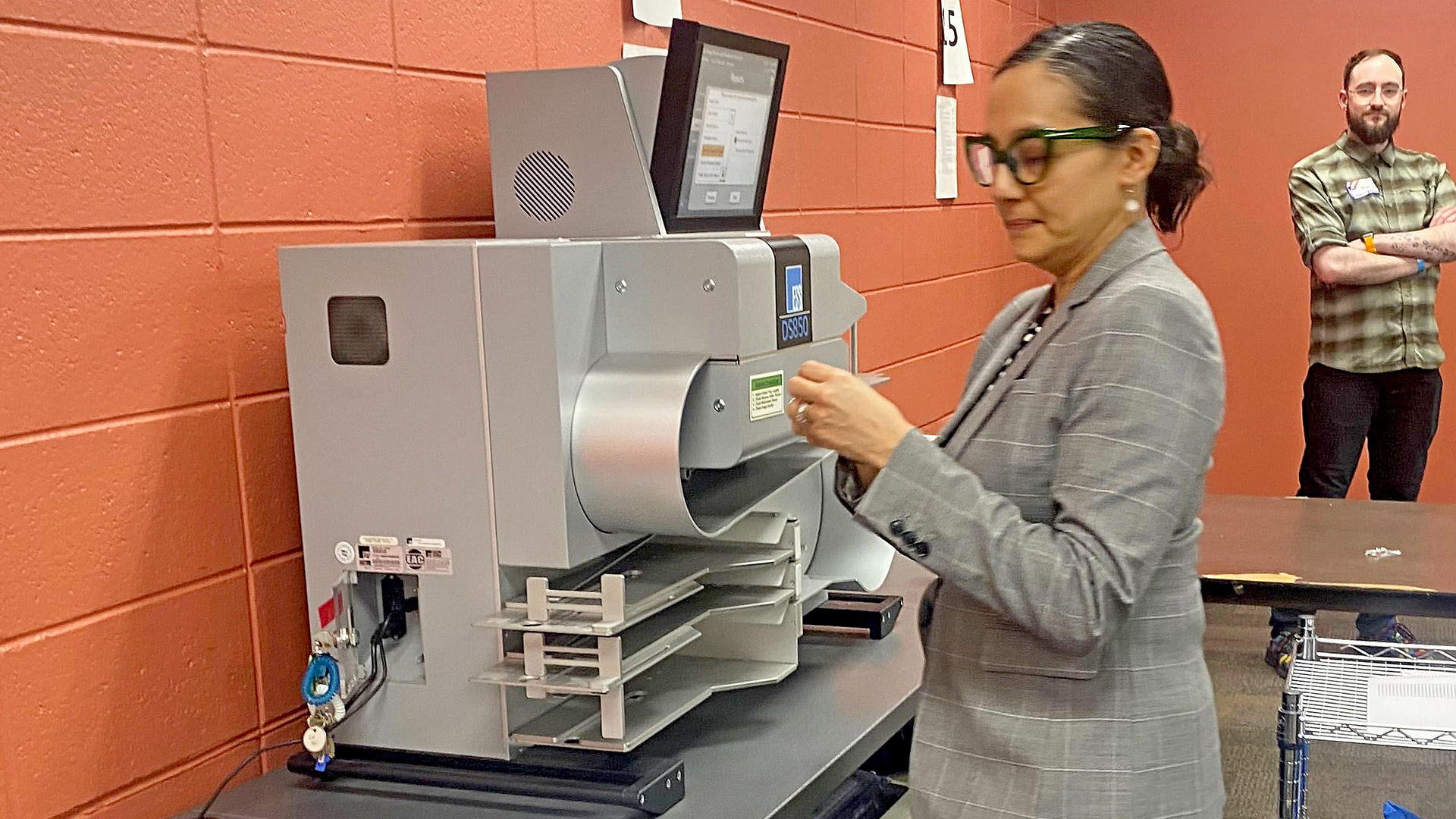 Paulina Gutierrez stands in front of a ballot tabulator machine placed on top a table, with another person standing in the background with crossed arms and wearing a disposable nametag, in a room with painted concrete block walls.
