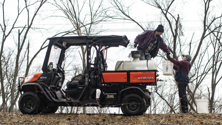One person stands next to a five-gallon plastic bucket and lifts another bucket and hands it to another person standing on the back of a utility task vehicle with a larger plastic tank in its cargo space, with the UTV parked on a ridgeline with leafless trees and power lines in the background.