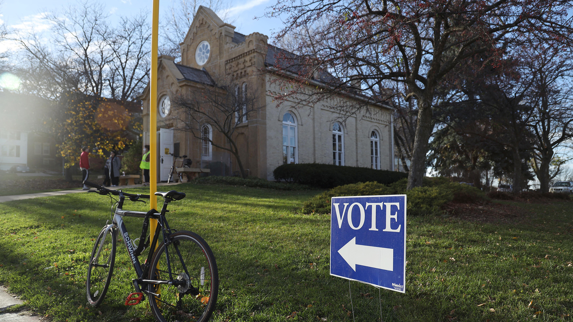 A cardboard sign with an arrow and the word "Vote" is mounted with metal poles on a lawn next to a bicycle locked to a street sign pole, in front of a stone masonry building with arched and rose windows with people standing outside its front entrance, with trees and buildings in the background under a party cloudy sky.