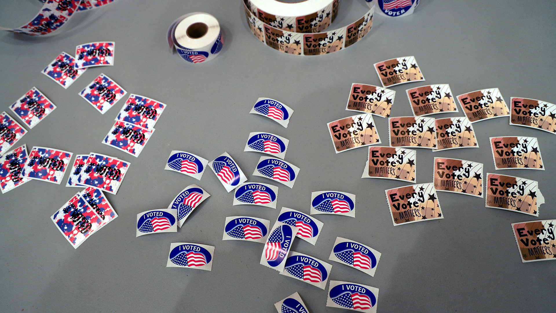 Three types of stickers — one showing a U.S. flag and the U.S. flag, another with an abstract design and the words "Every Vote Counts," and another with an abstract design and the words "Future Voter" — sit individually and in rolls on the surface of a table.
