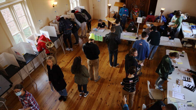 People sit at and stand in line facing multiple folding tables, with other people stand over temporary voting booths, in a room with wood floors and illuminated by daylight through windows and electric lights mounted to walls and on wood benches.