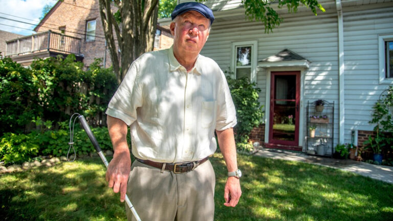 Don Natzke holds a white cane in his right hand while standing on a lawn outside a house with siding and a downspout next to a door, with a tree, foliage-covered fence and brick house in the background.
