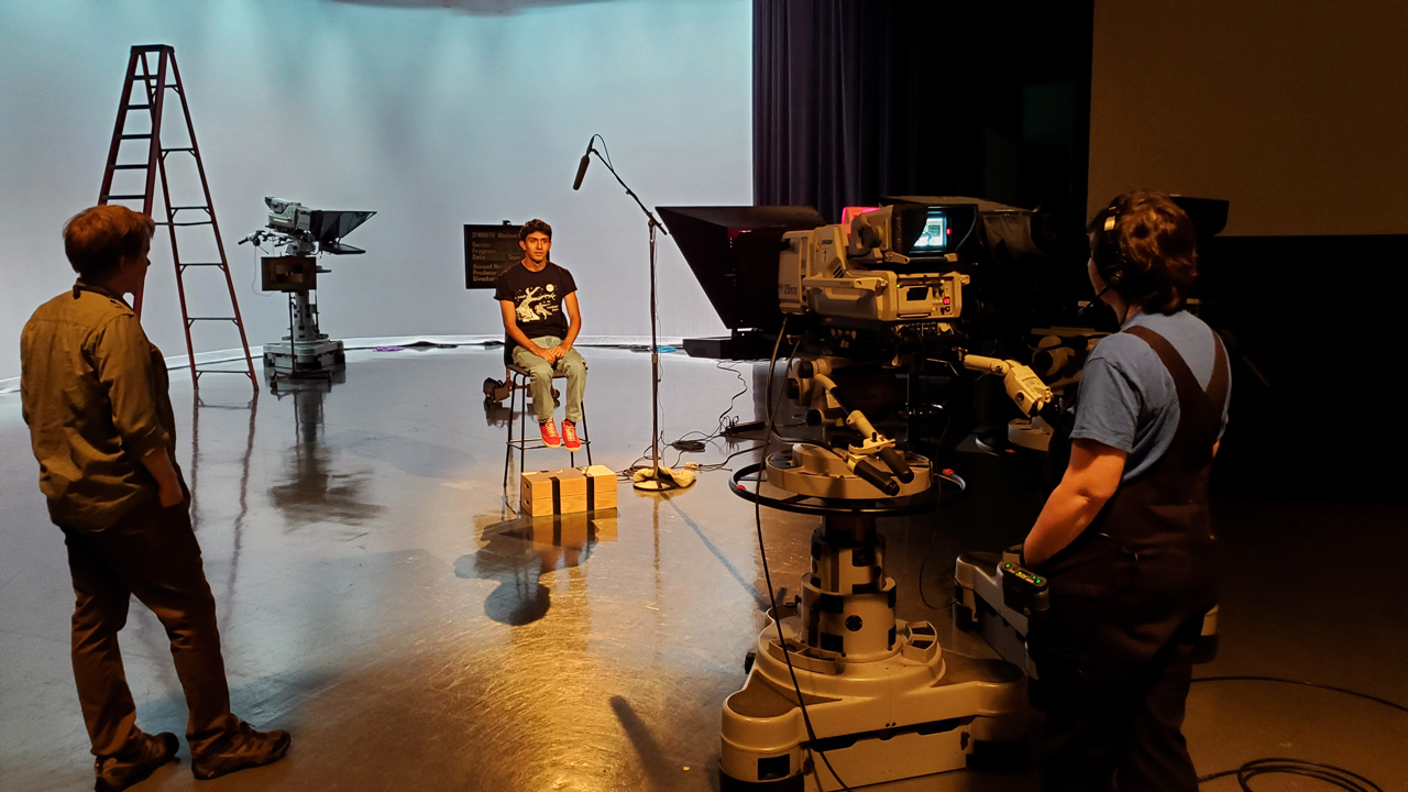A teenage boy sits in a chair for an interview in the PBS Wisconsin studio as a producer and operator stand behind the camera.