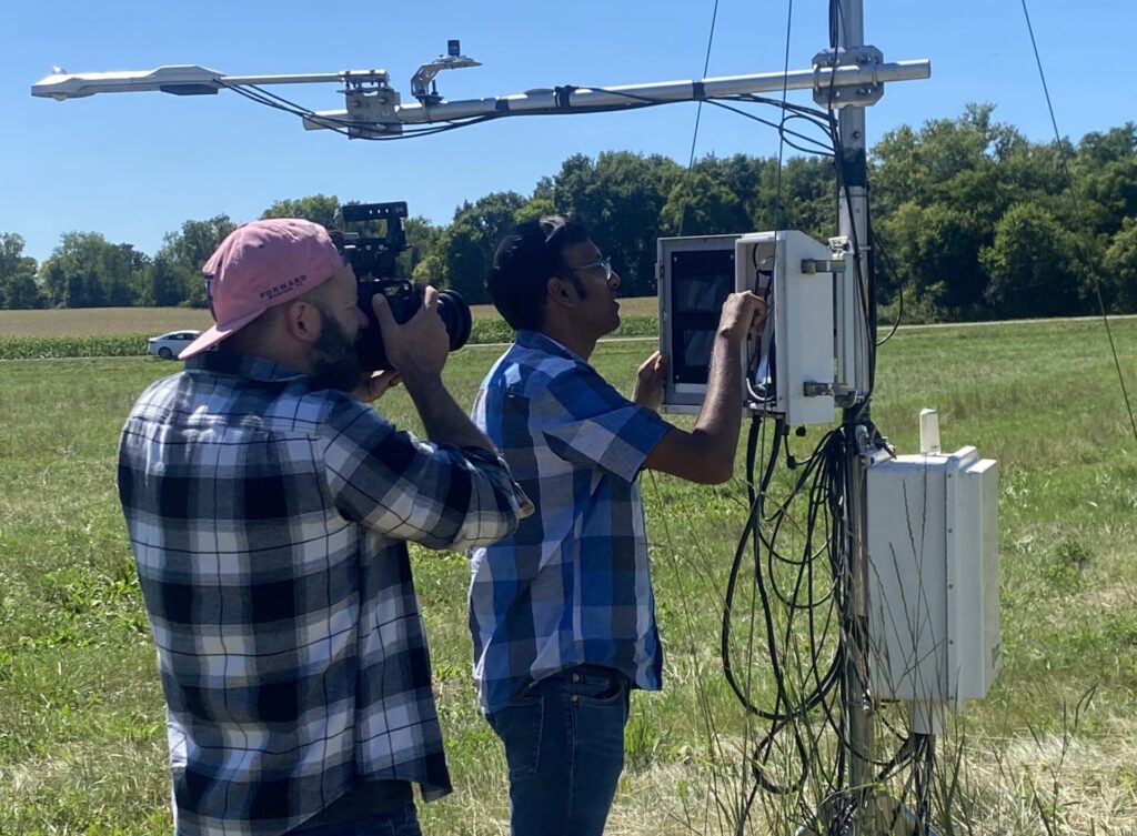Two people stand in a cattle field. One is taking measurements on a scientific instrument while the other person films.