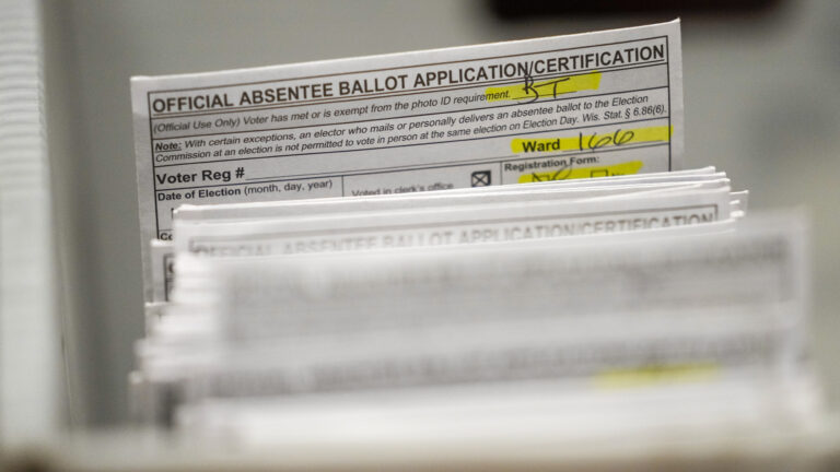 An envelope with the words Official Absentee Ballot Application/Certification at its top and multiple fields filed in with pens and highlighted with marker stands out among a stack of similar envelopes that are out-of-focus in the foreground.