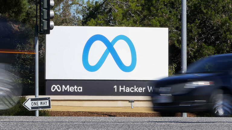 A sign showing a stylized infinity symbol above the same symbol next to the word Meta and the address 1 Hacker Way stands next to a road, with moving out-of-focus motor vehicles in the foreground and trees in the background.