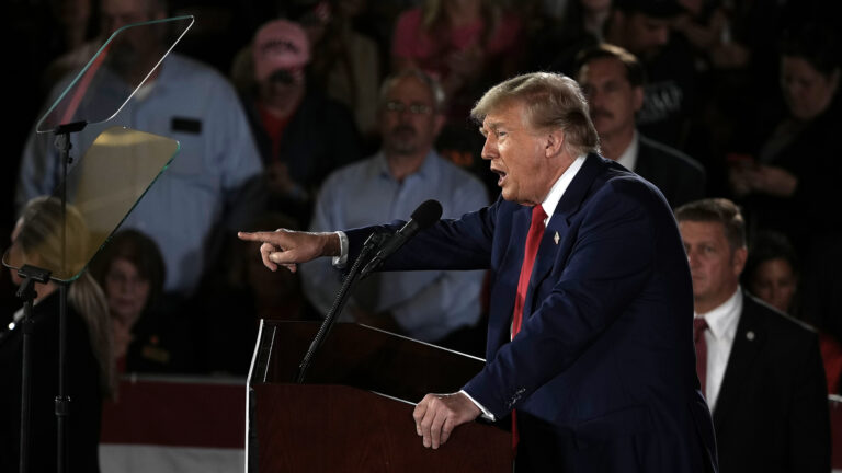 Donald Trump stands at a podium and holds its edge with his left hand and gestures with the index finger of his right hand while speaking into a microphone and facing teleprompter mirrors, with people standing in the background.