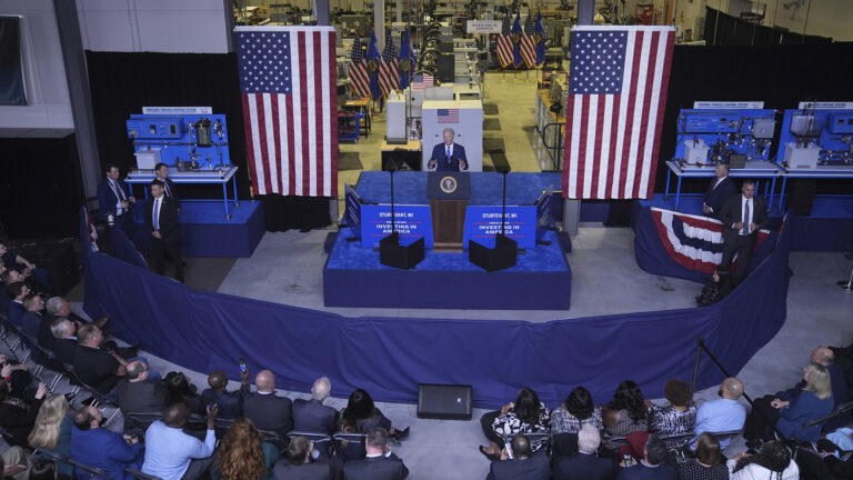 As seen from above, Joe Biden gestures with both hands and speaks while standing behind a podium with the Seal of the President of the United States affixed to its front, on a temporary riser with two teleprompters, with two large U.S. flags displayed on either side and several people standing behind a waist-high set of pipes and drapes separating the stage area from multiple rows of seated people facing toward him, with rows of U.S. and Wisconsin flags in the background of a large room with steel beams, shipping boxes and manufacturing equipment.