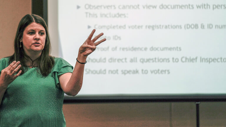 Claire Woodall stands and gestures with her hands while standing in front of a projection screen showing a list related to rules for election observers.