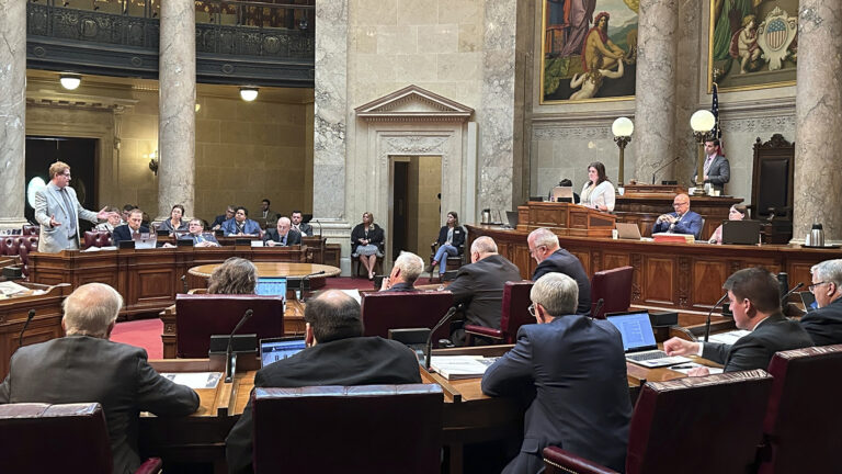 Tim Carpenter stands and gestures with both hands as other state senators sit at wood desks arranged in a semi-circle while other staff sit and stand at a wooden legislative dais, in a room with marble masonry and pillars, large paintings, and a second-story observation gallery.