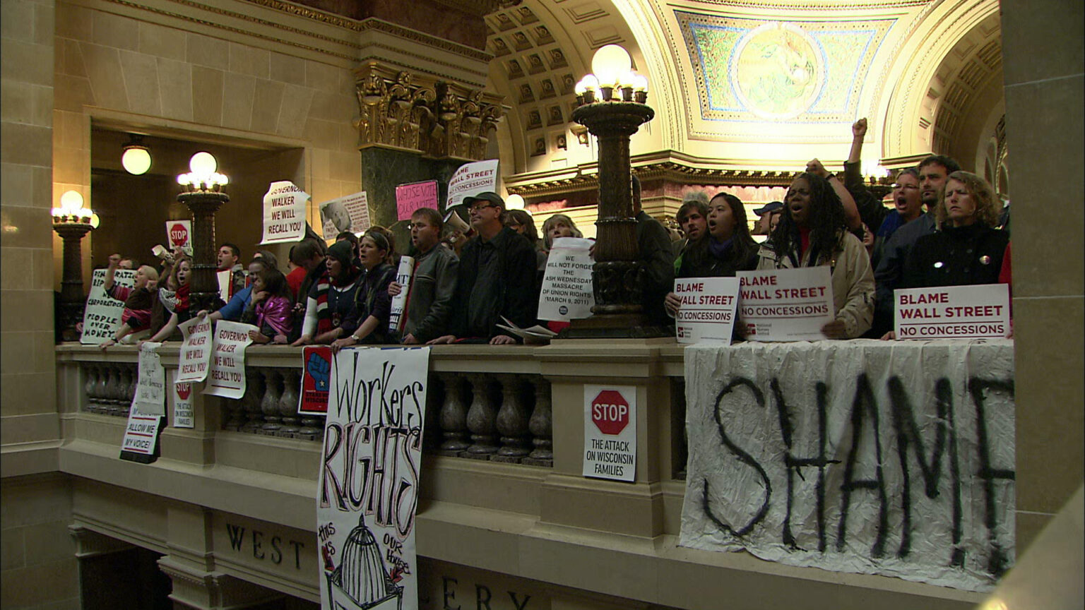 A group of people with signs like shame protest in the capitol.