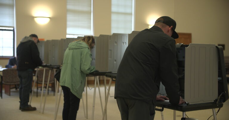 Three people stand at polls on election day 2023 and vote on paper ballots.