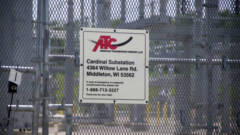 A sign showing the word mark for the American Transmission Company along with an address and emergency phone number is attached to a diamond-mesh metal fence, with out-of-focus electric power equipment in the background.