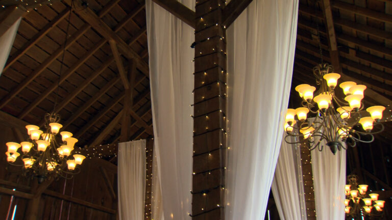 Metal chandeliers and long drapes hang from the rafters of the interior of a barn with wood-slat walls, with strings of electric lights wrapped around horizontal and vertical beams.