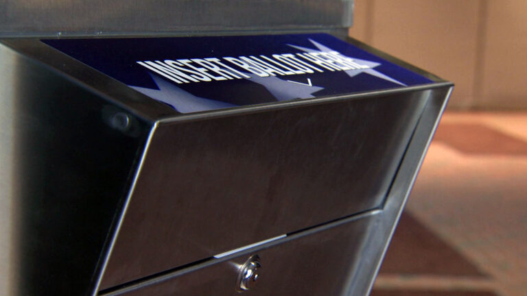 A metal box with a closed door, locking mechanism, and decal with star graphics and the words Insert Ballot Here stands in front of an out-of-focus wall.