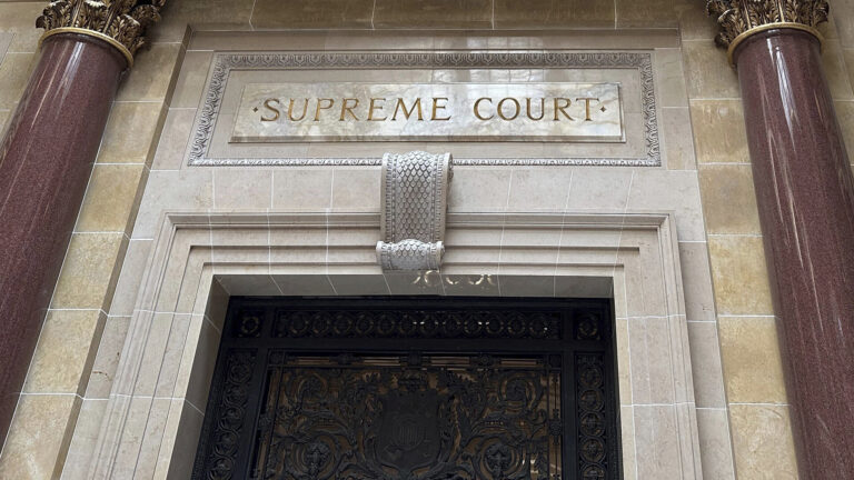 Marble pillars with Composite order capitals frame a doorway in a marble masonry wall with a carved sign reading Supreme Court above a metal filigree door.