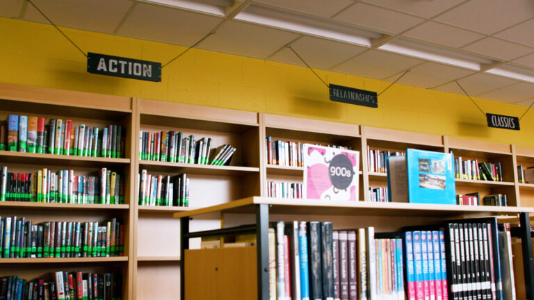 Books with library labels and binding fill tall wood bookcases that line one side of a room with painted concrete-block wall and overhead fluorescent lighting, with signs mounted on ceiling panels that read Action, Relationships and Classics, with a shorter wood and metal book case in the foreground.