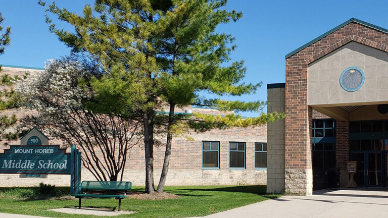 A wood sign with the address number 900 and the words Mount Horeb Middle Schools stands next to a metal park bench and in front of multiple trees on lawn between different wings of a masonry and brick building, with a sidewalk leading to glass doors at one entrance.
