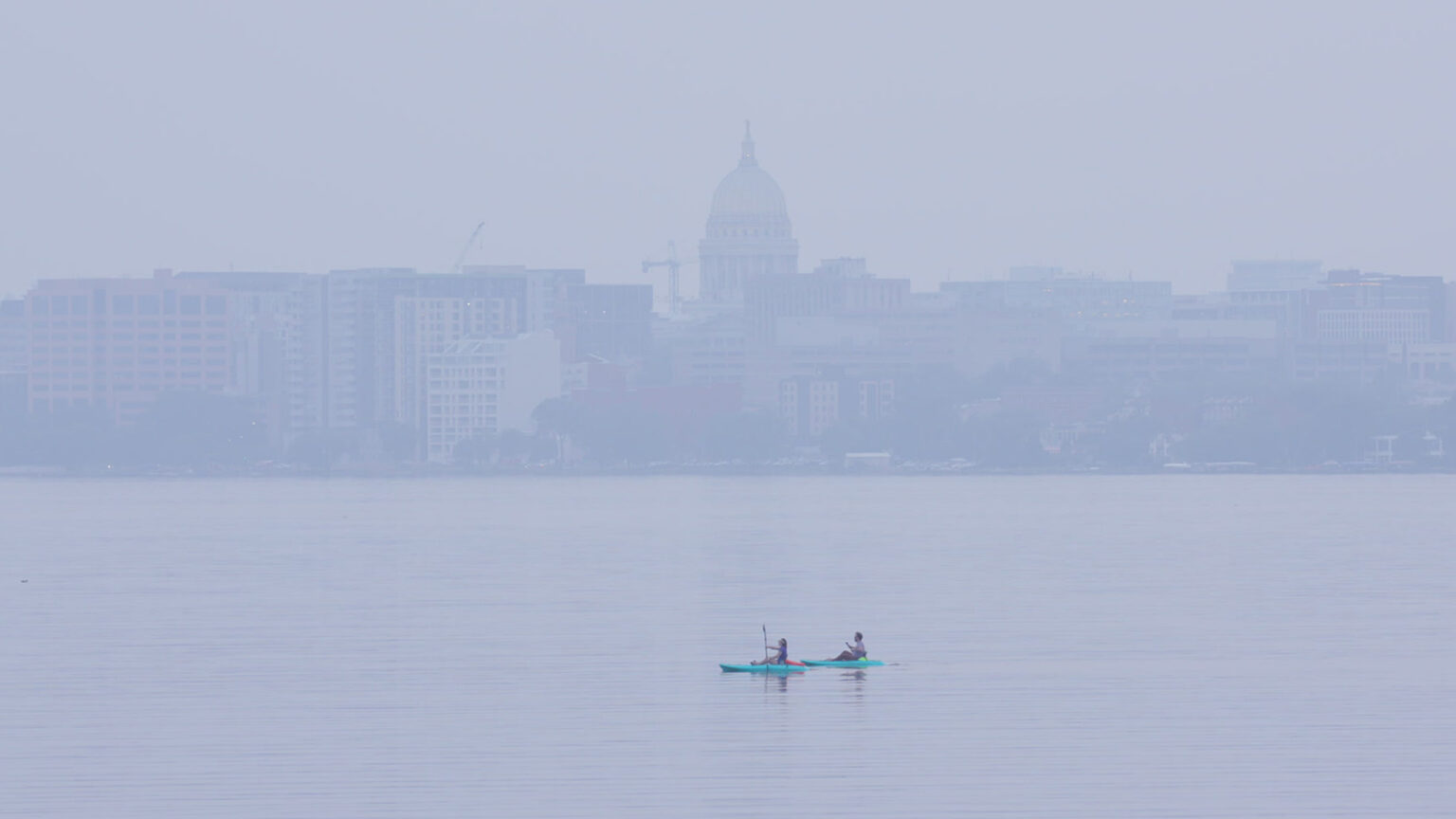 Two kayakers paddle in the calm waters of a lake, with the skyline of a city in the background with the Wisconsin State Capitol dome at center, with a haze from smoke obscuring the clarity of the buildings.