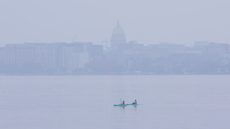 Two kayakers paddle in the calm waters of a lake, with the skyline of a city in the background with the Wisconsin State Capitol dome at center, with a haze from smoke obscuring the clarity of the buildings.