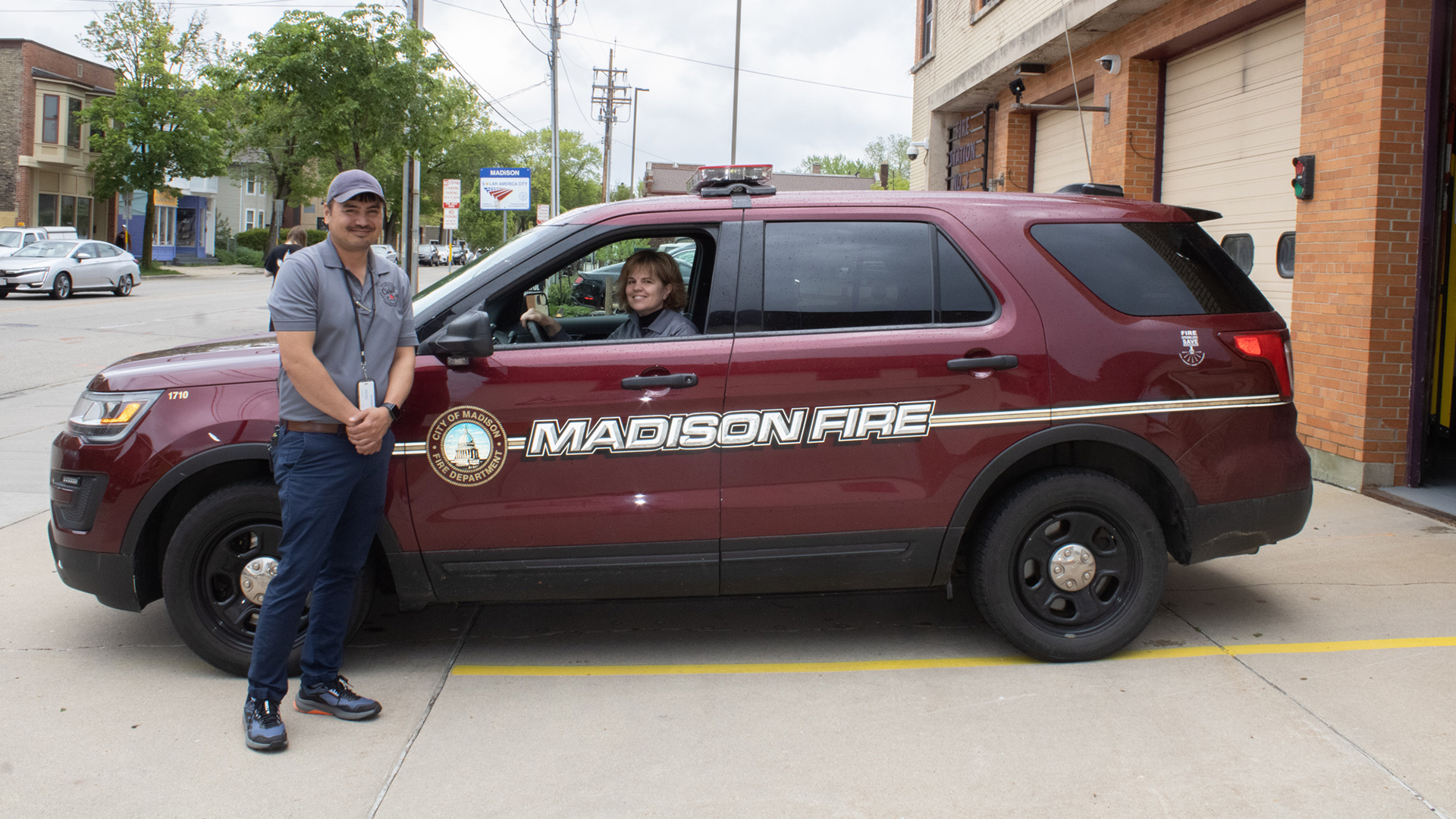 Amy Yelk Meinholz sits in the driver's seat of a red SUV with the City of Madison Fire Department logo and the words "Madison Fire" on its doors, while Eric Kinderman stands outside on a concrete driveway next to the driver's side mirror, outside a building with multiple garage doors with windows, with other vehicles, buildings and trees in the background.