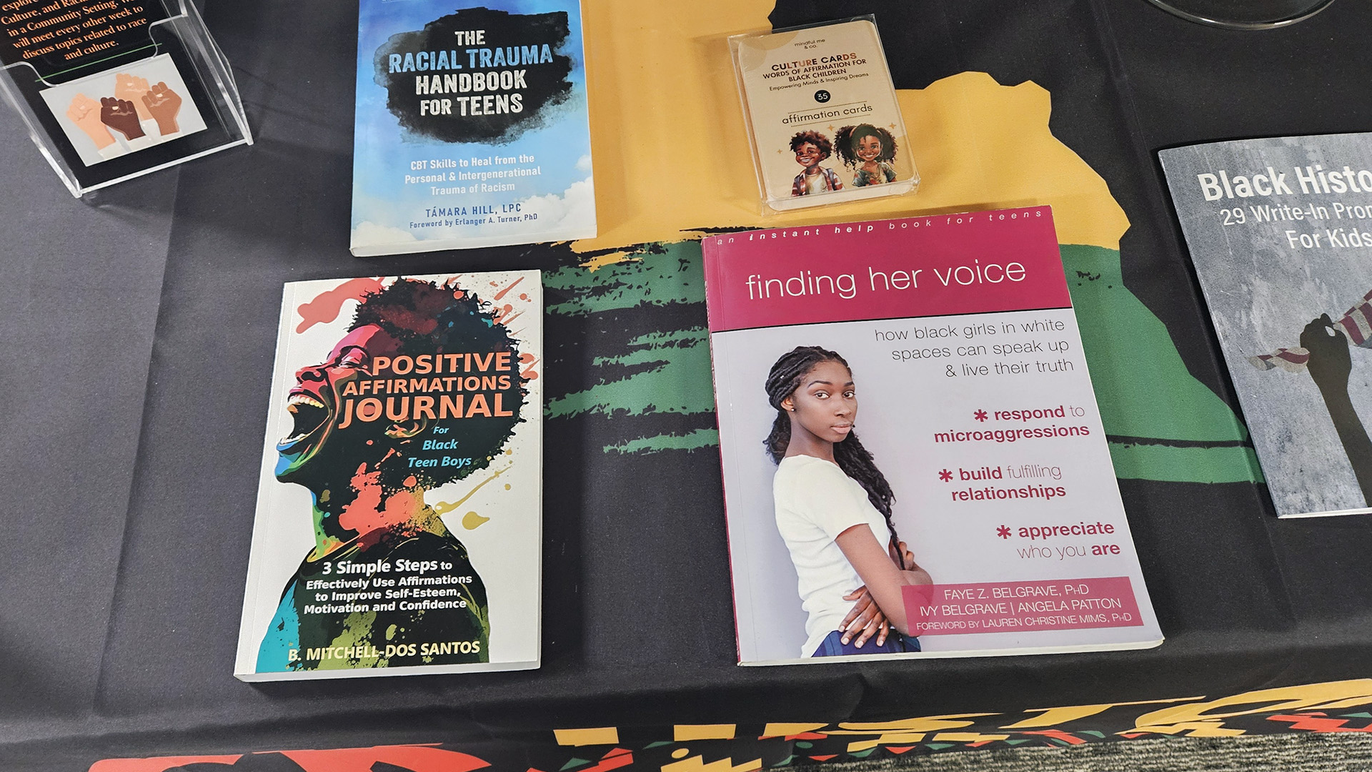 Multiple books, including those titled "The Racial Trauma Handbook for Teens," "Positive Affirmations Journal for Black Teen Boys," and "Finding Her Voice: How Black Girls in White Spaces Can Speak Up & Live Their Truth" sit atop a tablecloth with Pan-African colors on a table.