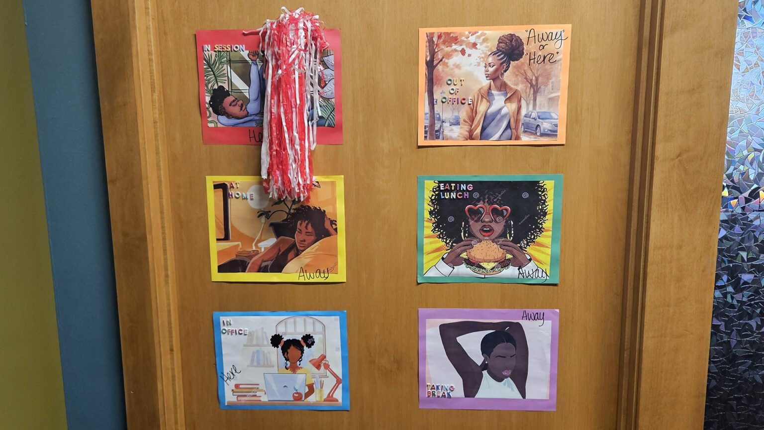 Six paper signs with illustrations of Black individuals engaged in different activities with the words In Session, Out of Office, At Home, Eating Lunch, In Office, and Taking Break, with a small pom pom attached to the one marked In Session, are attached to a wood door, with a painted wall on one side and a craquelure glass window on the other side.