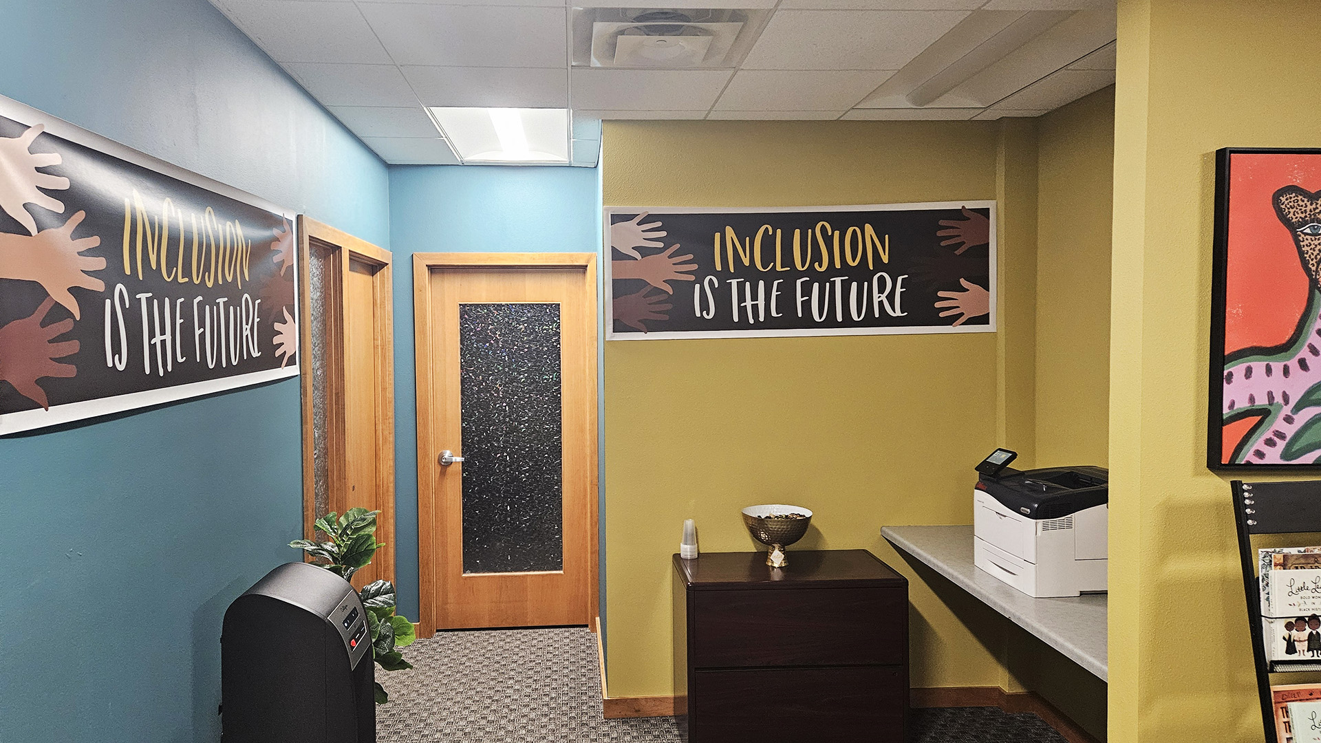 Vinyl signs with illustrated hands in different skin tones and with the words "Inclusion Is the Future" are attached to multiple walls of an office, with a printer on a wall-mounted table next to a wood stand with a bowl on its surface and multiple wood doors with craquelure glass windows.