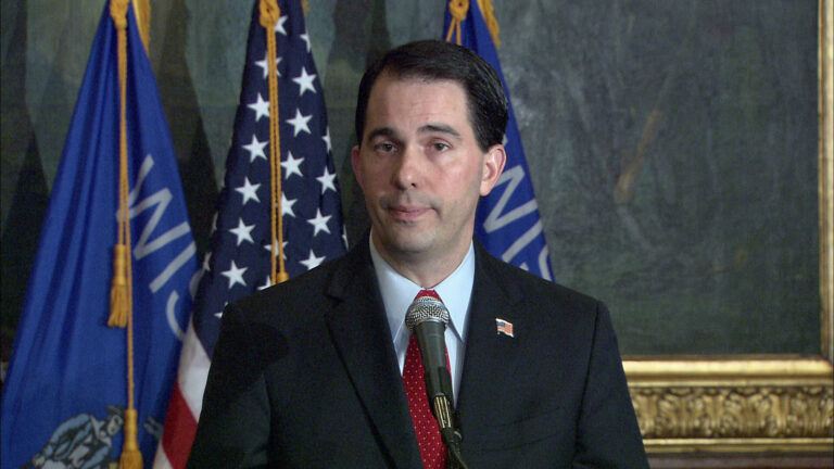 Scott Walker stands in the capitol in front of the state and American flags.