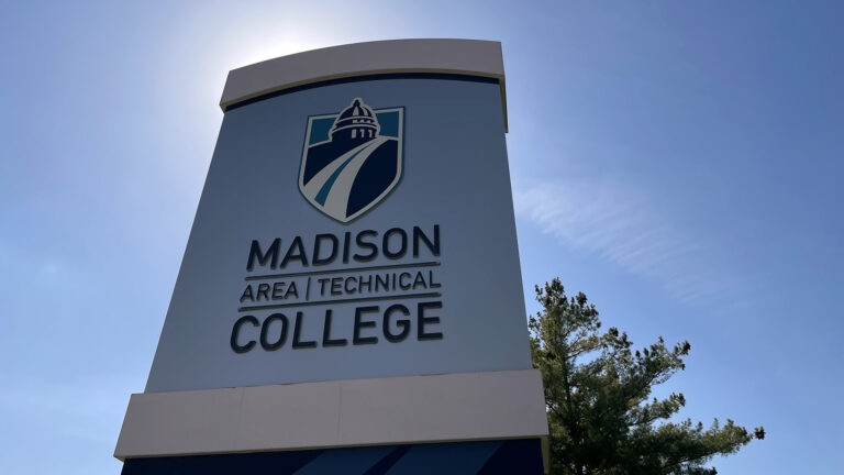 Sunlight illuminates the edges of a sign showing a shield-shaped stylized logo of a dome with a road leading to and from its base above the words Madison College in larger type and Area Technical in smaller type, with a tree and clear sky in the background.