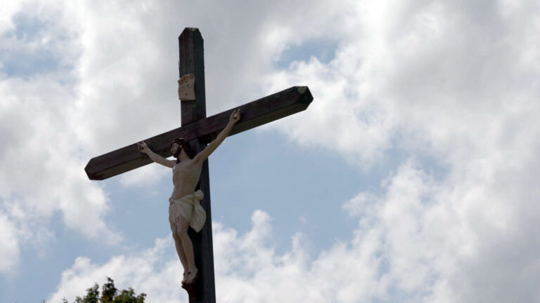 A wood crucifix with a painted figure of Jesus stands outdoors, with the top of a tree in the background under a partly cloudy sky.