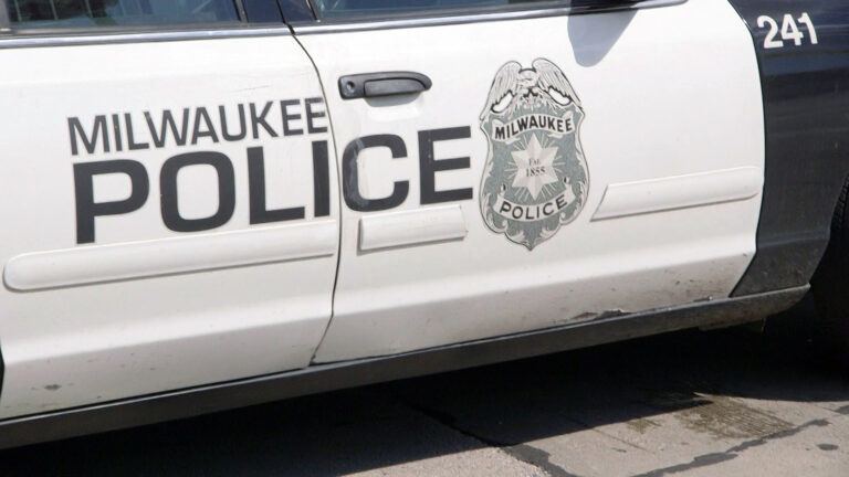 The badge-shaped logo of the Milwaukee Police Department, with a stylized eagle at its top and a six-pointed star with the words Est. 1855 in the middle of it, is painted on the side of parked black-and-white sedan between the words Milwaukee Police and the number 241.