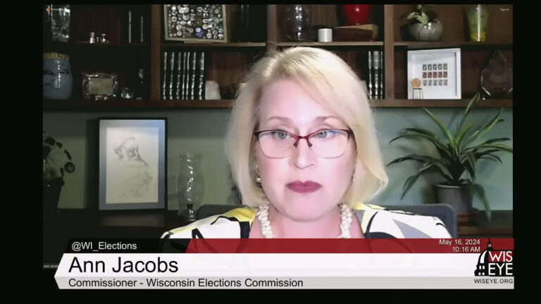 A video still image shows Ann Jacobs sitting in an office with wood shelves filled with books, plants and other items in the background, with a video graphic at bottom including the text Ann Jacobs and Commissioner - Wisconsin Elections Commission.