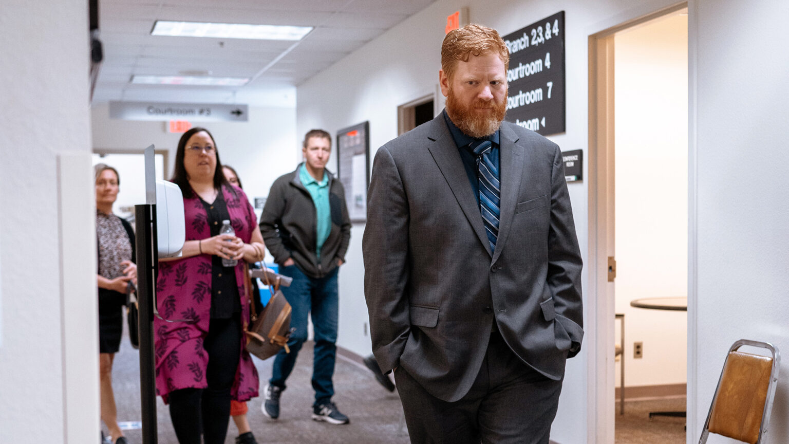 Jeremy Haney walks with his hands in his pockets with multiple people behind him in a hallway with signs noting courtroom numbers and fluorescent ceiling lights.