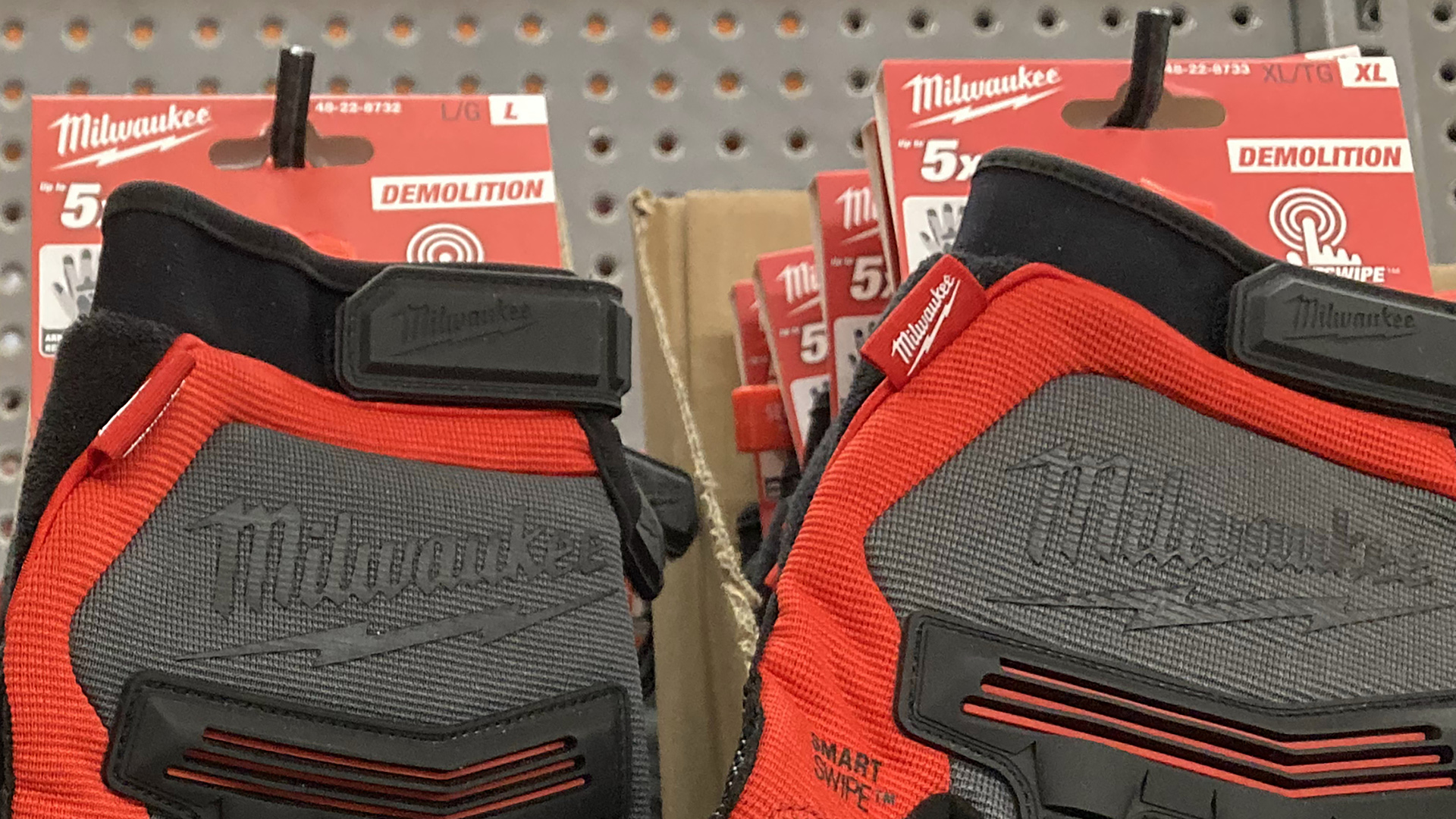 Two different sizes of work gloves with knuckle pads and Velcro wrist-fasteners are arranged on hangers on a shelf.
