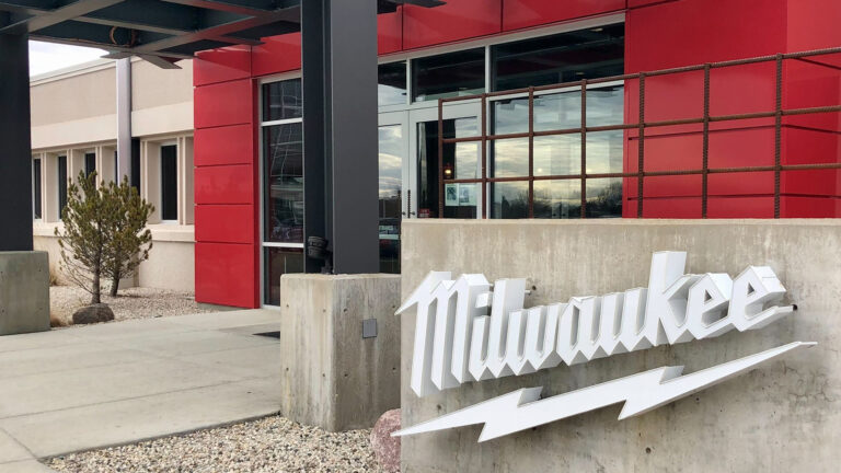 A sign with the Milwaukee Tool wordmark and lightning bolt logo stands next to the entrance of a building.