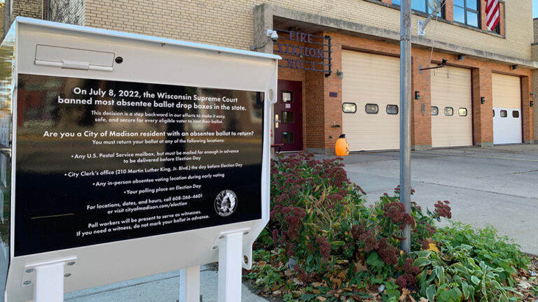 A metal container standing on four legs with a text graphics that starts with the line On July 8 2022, the Wisconsin Supreme Court banned most absentee ballot drop boxes in the state at its top stands on a concrete slab next to a garden plot, with a brick building with a metal sign reading Fire Station No. 3 and multiple garage doors in the background.
