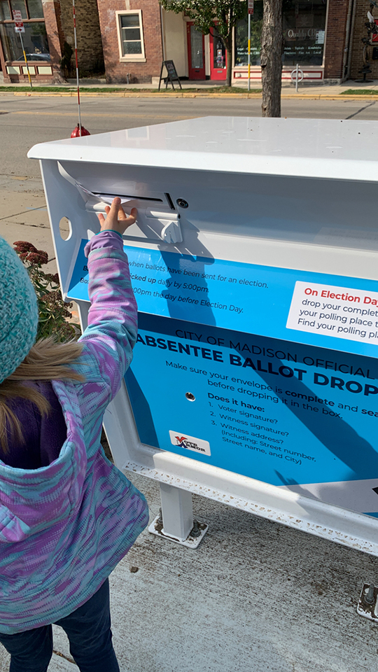 A child inserts a paper envelope into a narrow slot on the side of a large metal container with text graphics that include the words "City of Madison Official Absentee Ballot Drop Box" that is bolted to a concrete slab, with a street, trees and buildings in the background.