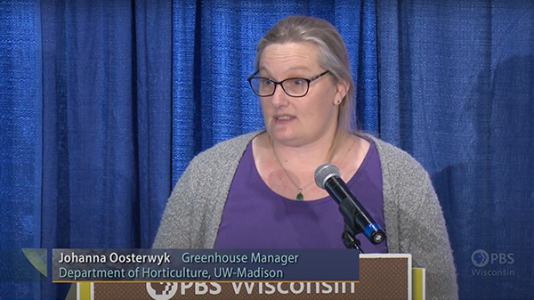 Johanna Oosterwyk, UW-Madison Horticulture Greenhouse Manager, presents during the Garden Expo from a brown podium in front of a blue background.