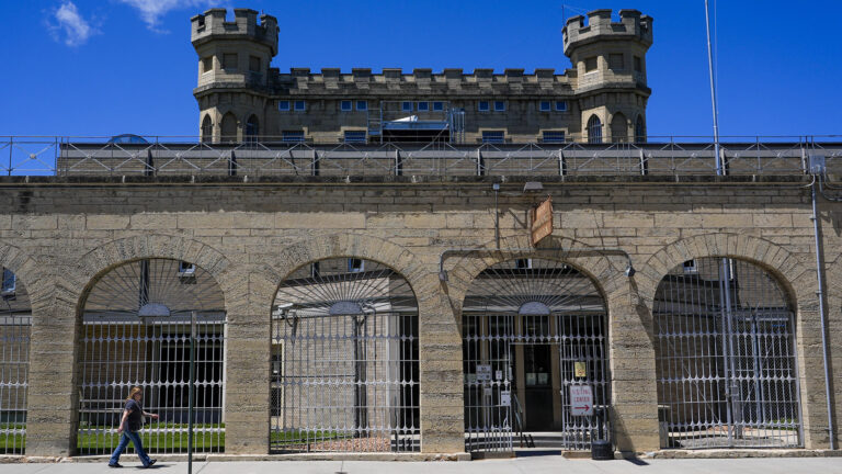 A person walks on a sidewalk toward a gate in a barred fence that stands open in a central archway of a masonry wall standing outside a masonry structure topped by two crenelated towers.