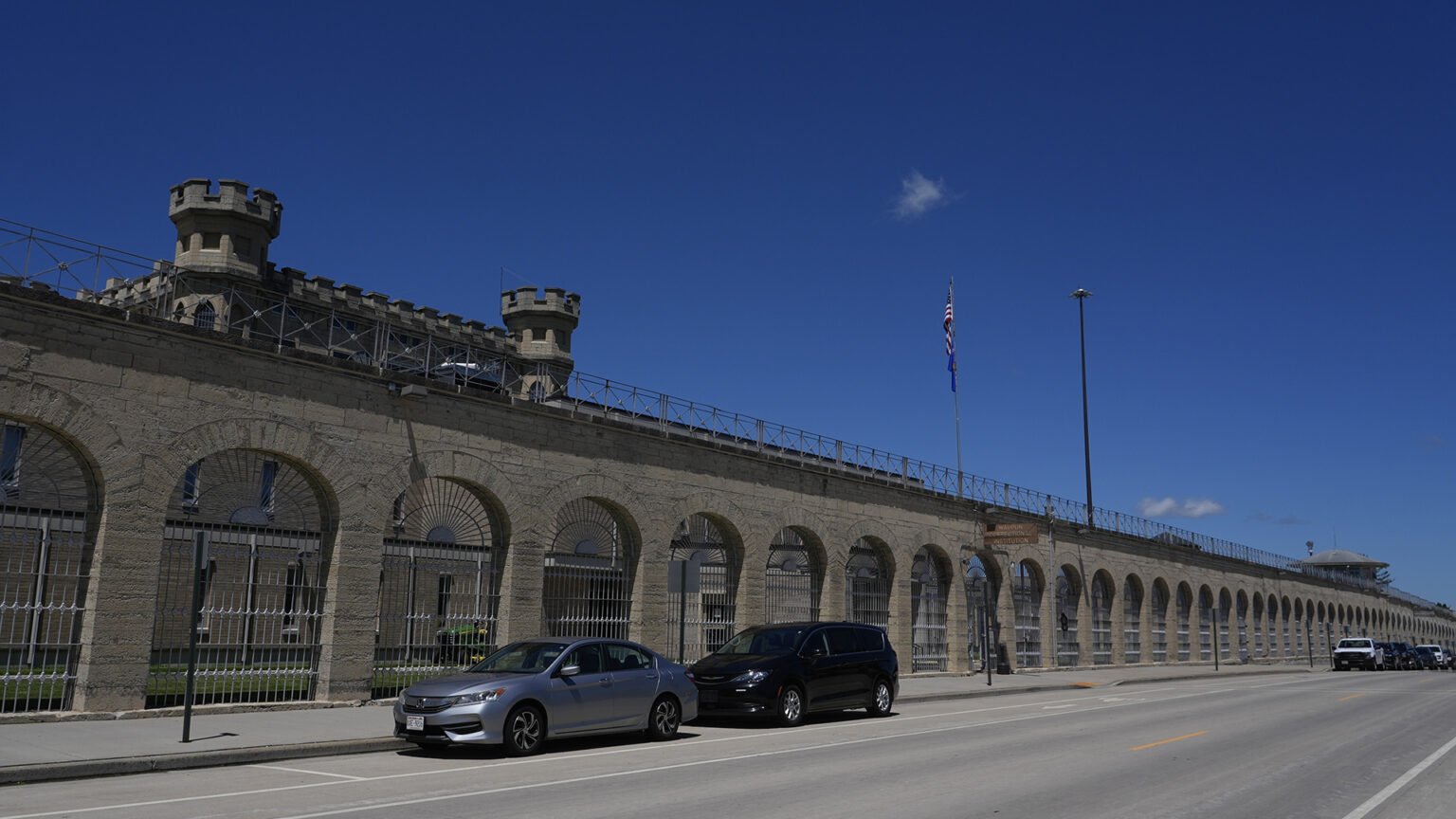 Multiple cars are parked in roadside parking spaces next to a masonry wall with numerous arches enclosed with a metal fence, which stands in front of a masonry structure topped by crenelated towers to one side and a tower with a peaked roof in the distance, along with a flagpole with the U.S. and Wisconsin flags and a tall light pole.