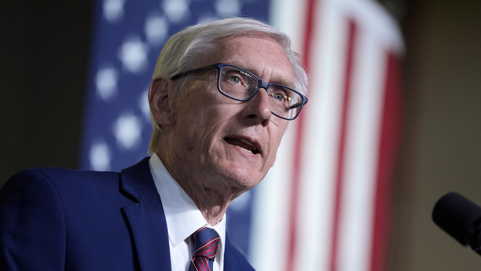 Tony Evers speaks into a microphone while standing in front of an out-of-focus U.S. flag.