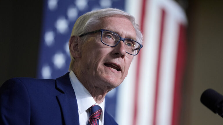 Tony Evers speaks into a microphone while standing in front of an out-of-focus U.S. flag.