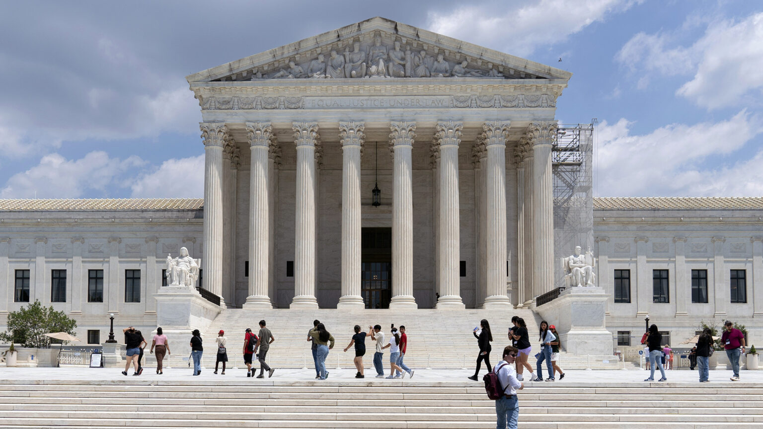 People stand on a steps and a plaza outside the west façade of the U.S. Supreme Court Building with marble masonry, a portico of columns flanked at their base by two sculptures of seated figures, and a pediment with a of sculptures of figures.
